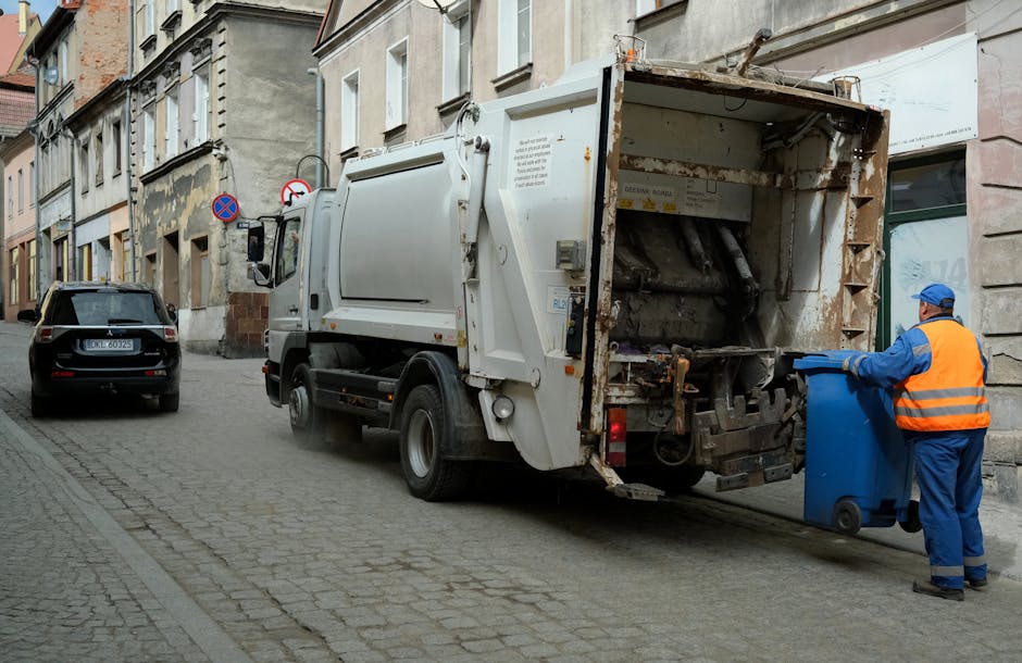 A street scene showing a white waste collection truck parked alongside a row of older, multi-story buildings with a rough, weathered facade. The rear of the truck is open, revealing rusted metal components and an empty, worn interior designed for rubbish compaction. A worker dressed in blue protective clothing and a high-visibility orange vest is in the process of disposing of a blue wheeled bin into the truck’s hopper, standing on a cobblestone pavement that extends along the street. Next to the truck, a black car is parked parallel to the kerb, with visible license plates and a sleek exterior finish. The environment appears to be a quiet urban area, with other signs such as a no-parking sign visible in the background. The overall scene captures an outdoor waste collection operation likely associated with private waste handling services, consistent with the context of reliable rubbish removal options.