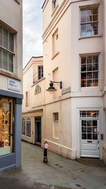 A narrow urban alleyway bordered by pale cream-colored, multi-story buildings with large sash windows and decorative architectural details. On the left, part of a shopfront is visible, including a display window with items and a sign indicating the address 'Avery Howe W1'. A vintage-style black wall-mounted lantern is affixed to the building in the center. The concrete pavement is clean, with a small bollard positioned near the alley's entrance, likely for traffic or pedestrian control. The scene is illuminated by natural daylight, with soft shadows cast on the building façades, suggesting a partly cloudy sky. The overall environment appears quiet and well-maintained, typical of a residential or mixed-use area in central London, where private waste collection and on-site clearance services, such as those provided by Rubbish Collection Mayfair, may be employed to manage refuse outside of local authority disposal options.