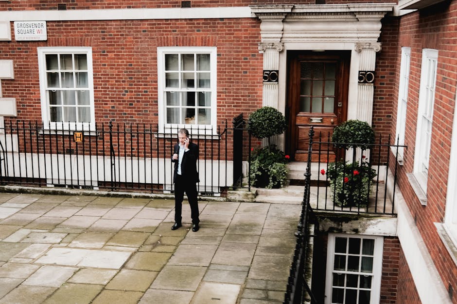 The image shows a man dressed in a black suit and tie standing on a paved sidewalk in front of a red-brick residential building with white-framed windows. He is holding a phone to his ear with his right hand and appears to be engaged in a conversation. In front of the building, there is a small, black metal fence enclosing a narrow garden area with two neatly trimmed, round green shrubs and some red flowering plants positioned beside the entrance door. The entrance features a wooden door with a glass panel, and the building's facade is decorated with stone architectural details around the door frame, including pilasters and an ornate entablature. A white wall-mounted mailbox is visible to the right of the door, and a street sign indicating 'Grosvenor Square W1' is affixed to the building to the upper left. The overall scene appears to be during daylight with natural, even lighting, and the setting suggests an urban residential area focused on private property and independent waste collection services occasionally seen in the vicinity.