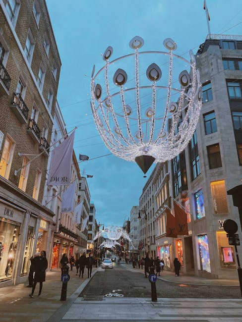 A narrow urban alleyway bordered by pale cream-colored, multi-story buildings with large sash windows and decorative architectural details. On the left, part of a shopfront is visible, including a display window with items and a sign indicating the address 'Avery Howe W1'. A vintage-style black wall-mounted lantern is affixed to the building in the center. The concrete pavement is clean, with a small bollard positioned near the alley's entrance, likely for traffic or pedestrian control. The scene is illuminated by natural daylight, with soft shadows cast on the building façades, suggesting a partly cloudy sky. The overall environment appears quiet and well-maintained, typical of a residential or mixed-use area in central London, where private waste collection and on-site clearance services, such as those provided by Rubbish Collection Mayfair, may be employed to manage refuse outside of local authority disposal options.