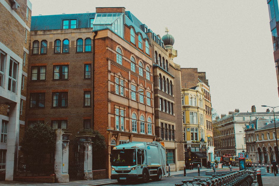 A city street scene featuring a range of historic and modern buildings, including a prominent multi-story brick structure with decorative arched windows and a copper-green domed tower at the top. To the right, there is a pale yellow building with intricate architectural details and large sash windows, complemented by other nearby structures with varied facades. In the foreground, a light blue waste collection truck with the company name 'Rubbish Collection Mayfair' is parked along the curb amid bollards and bicycle racks. The street is lined with asphalt, and a few pedestrians and additional vehicles are visible in the background. Overcast sky provides diffuse lighting, highlighting the textures and colors of the buildings and street scene, which illustrates an environment suitable for private waste collection or on-site clearance services in a busy urban setting.