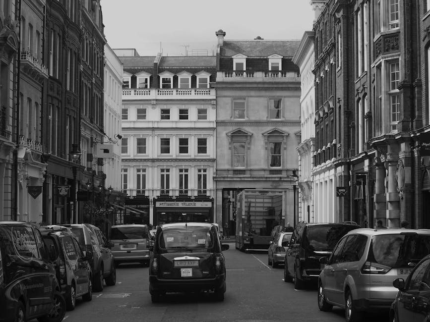 A city street scene featuring a range of historic and modern buildings, including a prominent multi-story brick structure with decorative arched windows and a copper-green domed tower at the top. To the right, there is a pale yellow building with intricate architectural details and large sash windows, complemented by other nearby structures with varied facades. In the foreground, a light blue waste collection truck with the company name 'Rubbish Collection Mayfair' is parked along the curb amid bollards and bicycle racks. The street is lined with asphalt, and a few pedestrians and additional vehicles are visible in the background. Overcast sky provides diffuse lighting, highlighting the textures and colors of the buildings and street scene, which illustrates an environment suitable for private waste collection or on-site clearance services in a busy urban setting.