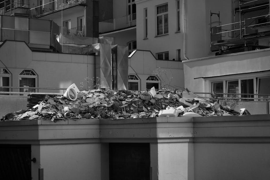 A large pile of demolition debris and rubble sits atop a flat rooftop or terrace in an urban area, with various materials including broken bricks, concrete fragments, shattered plaster, and discarded insulation visible. The debris is unevenly spread across the surface, with some pieces leaning against each other and others scattered loosely. Among the debris, there are white plastic or styrofoam objects, possibly insulation or packaging, partially embedded within the rubble. The building structures surrounding the site feature modern architectural elements with light-colored facades, rectangular windows with decorative borders, and some sections of scaffolding or external construction equipment visible on the right. The background includes multi-storey residential or commercial buildings, indicating a densely built city environment. The lighting appears to be natural daylight, casting subtle shadows and highlighting the texture and arrangement of the debris, suggesting an on-site clearance process that could be handled by private waste removal services such as Rubbish Collection Mayfair, specializing in rubbish or demolition debris disposal in central London premises.