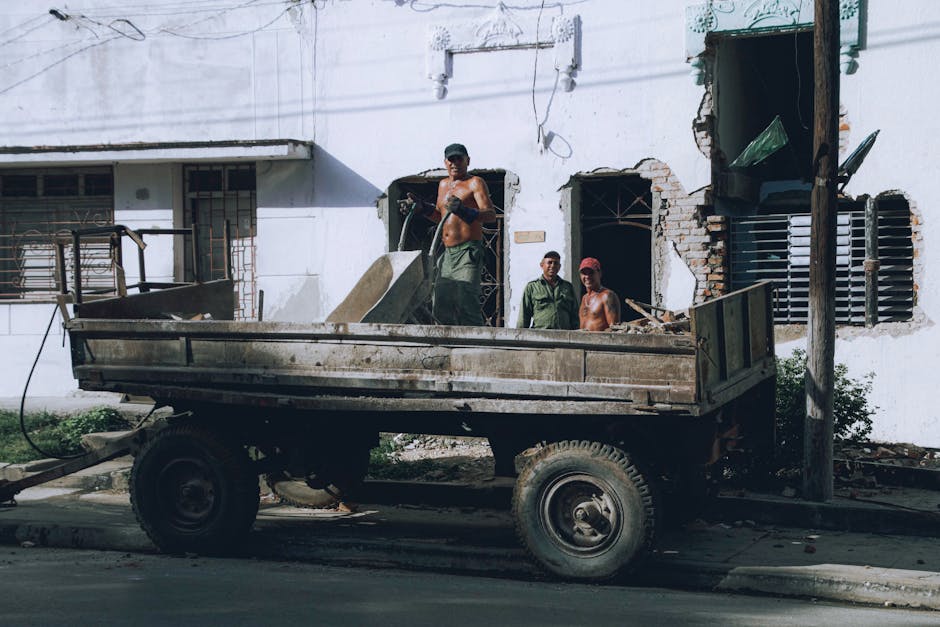 A group of three workers standing on the back of an old, rusty flatbed truck is depicted in this image, positioned beside a building with a white, partially damaged exterior wall. One worker, shirtless and wearing dark trousers and a cap, is lifting or adjusting a large, flat piece of debris that appears to be construction material. The second worker, dressed in casual clothing with a cap, stands behind him, observing, while the third worker, also shirtless and wearing a cap, leans against the building's wall, near the open doorway. The building’s exterior shows signs of demolition or renovation, with bricks, concrete, and loose debris visible around the doorway and window. The flatbed truck is parked on a street with visible pavement, and the scene is illuminated by natural daylight, highlighting the construction or renovation activity in an urban setting, which suggests a site of private waste clearance or on-site debris removal. Rubbish Collection Mayfair offers similar private rubbish removal services for local renovation waste disposal.