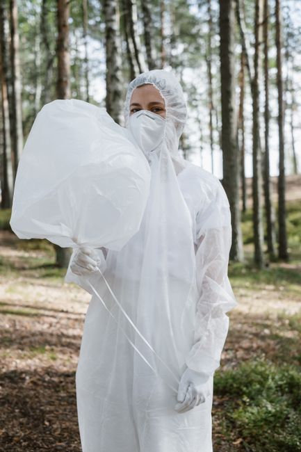 A person dressed in full white protective personal equipment, including a hooded coverall suit, gloves, and a face mask, stands outdoors in a forested area with tall, slender trees and green foliage in the background. The individual is holding a large, white, semi-transparent plastic bag filled with waste, gripping it by the top with one hand while the bag appears to be slightly expanded and irregular in shape, suggesting it contains rubbish. The background shows diffused natural light filtering through the tree canopy, creating a soft, natural atmosphere typical of woodland environments. The scene relates to waste collection or disposal, possibly indicating an outdoor rubbish removal service such as independent waste handling or environmental cleanup, with the individual prepared for careful and safe rubbish collection in an outdoor setting. This visual aligns with services like those provided by Rubbish Collection Mayfair, highlighting the process of on-site waste management in non-urban areas, where specialised personal protective equipment is used for safe handling of rubbish in environmentally sensitive locations.