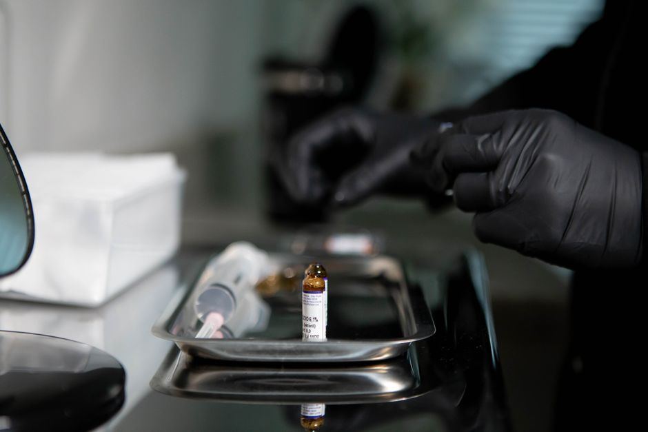 A healthcare professional wearing black nitrile gloves is handling medical vials and syringes on a stainless steel tray, situated on a sterile laboratory or clinical setting surface. The scene includes a syringe with a clear barrel and pink plunger, and a small amber glass vial with a white label displaying text, both placed on the reflective tray. In the background, there is a blurred white box and a partially visible medical device. The environment is lit with neutral, subdued lighting that contributes to a clinical atmosphere, emphasizing cleanliness and safety. The detailed textures of the gloves, the smooth surface of the tray, and the clear and amber materials of the medical items are all distinctly visible. The image subtly relates to medical waste or hazardous waste disposal, aligning with independent or private waste handling practices, and may support content about safe collection and disposal of clinical waste by specialists like Rubbish Collection Mayfair, without assuming specific disposal methods.