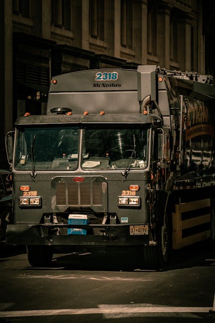 A large commercial rubbish collection truck, painted in dark green and grey, facing directly forward on a city street at night. The truck features a prominent front grille with a small, oval-shaped emblem in the centre, and has multiple orange warning lights on the top and sides. The vehicle's windshield is expansive, showing the dashboard and steering wheel inside, with various papers or items visible on the dashboard. The vehicle's identification number '2318' is displayed in blue above the windshield and on the upper corners of the cab, with the brand name 'McNeilus' printed in black just below the number. The background includes a portion of a multi-storey building with stone or concrete facades, illuminated by street lighting, creating a slightly reflective glow on the polished surface of the truck. The scene suggests an urban environment where independent waste collection, perhaps by a private company like Rubbish Collection Mayfair, is undertaken as part of a commercial waste management service, emphasizing on-site clearance and alternative waste handling solutions in a city setting at night.