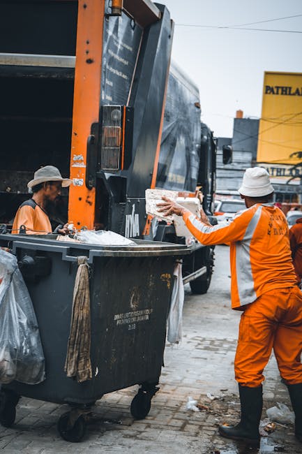 In an outdoor urban setting, two workers in bright orange high-visibility uniforms and white hats are engaged in waste collection near a large black rubbish bin mounted on a vehicle. One worker is seen extending their arm to place a piece of rubbish, possibly a paper or cardboard item, into or onto the bin, which is situated close to the side of a large, dark grey refuse truck equipped with side loading mechanisms. The scene captures a moment of active waste collection, with the worker's gloves visible as they handle the waste. The ground beneath them consists of paved bricks, and in the background, there are other vehicles, industrial buildings, and a yellow sign, indicating a busy urban environment. The lighting is natural, with no harsh shadows, suggesting a typical day with overcast or neutral lighting conditions, emphasizing the practical and professional nature of the waste management operation performed by Rubbish Collection Mayfair in a setting where private or alternative waste handling methods are being utilized in the cityscape.