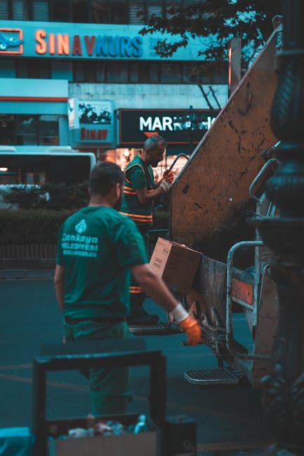 A large commercial rubbish collection truck, painted in dark green and grey, facing directly forward on a city street at night. The truck features a prominent front grille with a small, oval-shaped emblem in the centre, and has multiple orange warning lights on the top and sides. The vehicle's windshield is expansive, showing the dashboard and steering wheel inside, with various papers or items visible on the dashboard. The vehicle's identification number '2318' is displayed in blue above the windshield and on the upper corners of the cab, with the brand name 'McNeilus' printed in black just below the number. The background includes a portion of a multi-storey building with stone or concrete facades, illuminated by street lighting, creating a slightly reflective glow on the polished surface of the truck. The scene suggests an urban environment where independent waste collection, perhaps by a private company like Rubbish Collection Mayfair, is undertaken as part of a commercial waste management service, emphasizing on-site clearance and alternative waste handling solutions in a city setting at night.