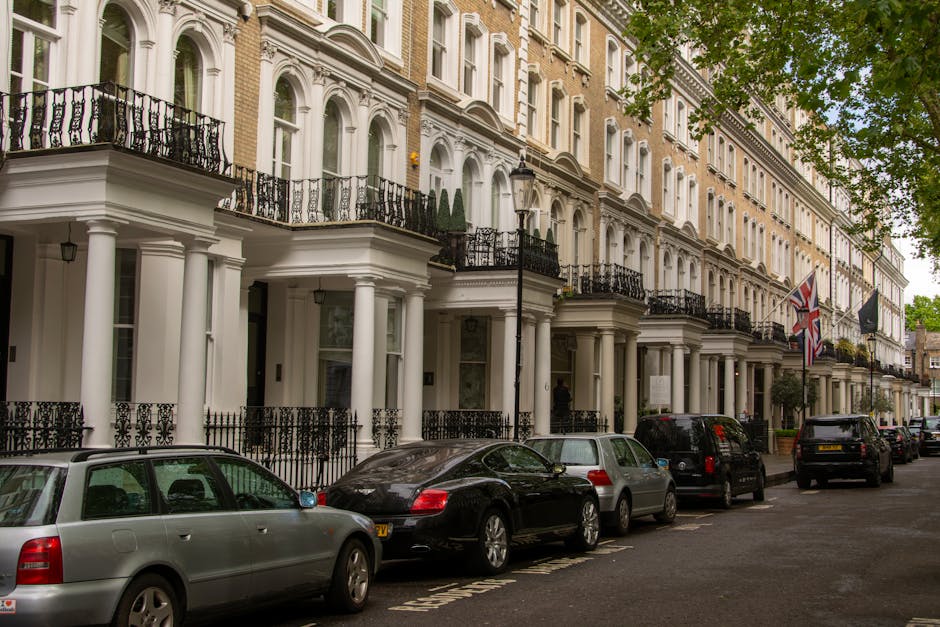 A row of white, multi-story Victorian-style townhouses with decorative columns, black wrought iron railings on small front balconies, and large sash windows, located along a tree-lined street. Several cars, including a silver station wagon, a black coupe, and a silver hatchback, are parked parallel to the curb in the foreground, with additional vehicles further down the street. The scene is captured during daytime under partly cloudy skies, with green leaves visible on the trees extending over the sidewalk. An ornate black street lamp stands near the center, and British flags are displayed on the building facades, indicating a typical urban setting that may benefit from private rubbish removal services. The overall atmosphere suggests a well-maintained residential area where occasional rubbish clearance or on-site waste collection could be part of property management activities, supported by local waste specialists such as Rubbish Collection Mayfair.