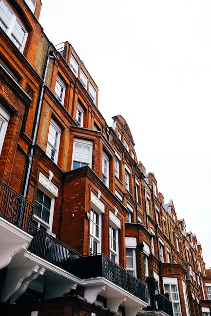 A multi-storey red-brick residential building situated along a city street, featuring large white-framed sash windows and decorative white stone accents above some windows. The building's facade includes multiple protruding bay windows and ornate cornices, with wrought iron railings on small balconies visible on several floors. The ground floor has a white architectural detail supporting a small balcony, while the neighbouring structures share similar brickwork and window designs. The sky above is overcast, casting a diffuse light on the scene, and the surroundings suggest an urban environment with closely spaced buildings characteristic of a cityscape. The building's exterior appears well-maintained, with the brickwork clean and intact, overlooking a quiet street where private property and potential opportunities for on-site waste clearance could be relevant for rubbish removal services such as those offered by Rubbish Collection Mayfair, especially in the context of house clearance or refurbishment projects.