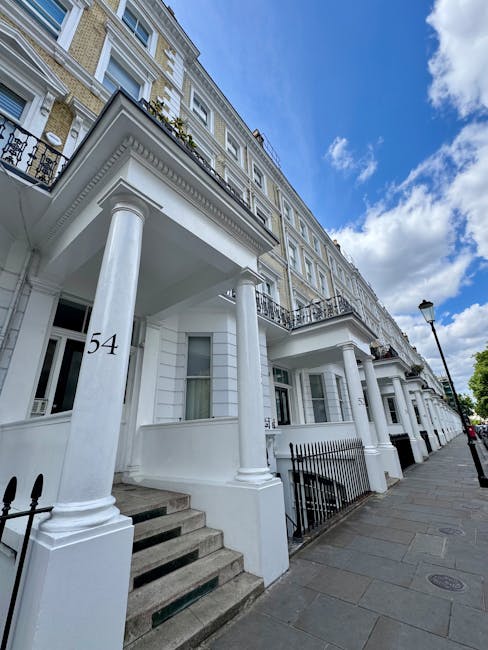 A row of white, multi-story Victorian-style townhouses with decorative columns, black wrought iron railings on small front balconies, and large sash windows, located along a tree-lined street. Several cars, including a silver station wagon, a black coupe, and a silver hatchback, are parked parallel to the curb in the foreground, with additional vehicles further down the street. The scene is captured during daytime under partly cloudy skies, with green leaves visible on the trees extending over the sidewalk. An ornate black street lamp stands near the center, and British flags are displayed on the building facades, indicating a typical urban setting that may benefit from private rubbish removal services. The overall atmosphere suggests a well-maintained residential area where occasional rubbish clearance or on-site waste collection could be part of property management activities, supported by local waste specialists such as Rubbish Collection Mayfair.
