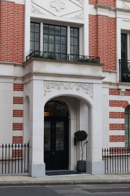 The image shows the front entrance of a multi-story building with a white stucco and red brick facade. The entrance features a large black door with a decorative metal grille, set within an arched white stone surround. Above the door, there is a small balcony with a black wrought iron railing, adorned with potted plants and green foliage. To the right of the entrance, a second window with black trim and clear glass is visible, framed by brickwork with a white stone trim around the window arch. The sidewalk directly in front of the entrance is paved, and a small black metal fence borders the property, with a neatly trimmed, potted topiary tree positioned on the right side of the door, inside a grey pot. The lighting appears natural, suggesting daylight, and the building’s exterior showcases a combination of textured brick and smooth stone finishes, typical of urban properties that may require occasional waste removal or on-site clearance services, as provided by Rubbish Collection Mayfair.