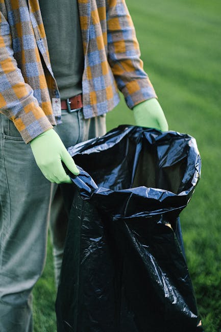 A person standing outdoors on a grassy area during daylight, wearing a checkered shirt and grey trousers, is visible from the waist down. They are wearing light green gloves and holding open a black plastic rubbish bag, which is partially filled with waste. The background features lush green grass and a slightly blurred natural environment, indicating an outdoor setting suitable for waste collection or environmental cleanup activities. The focus is on the individual’s hands and the rubbish bag, suggesting waste disposal or collection work, which aligns with private rubbish removal services. The image emphasizes careful handling and disposal of waste materials in an organized manner, consistent with professional rubbish collection practices.