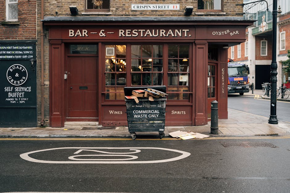 In front of a brick-fronted restaurant with a deep red wooden facade, a large black waste bin labeled 'Commercial Waste Only' is positioned on the pavement. The bin is filled with flattened cardboard boxes and paper waste, with some pieces spilling out onto the sidewalk nearby. To the left of the bin, a small chalkboard sign advertises a self-service buffet, and a street sign above indicates 'Crispin Street' at the corner. On the right side, a black bollard and a metal post are situated on the sidewalk, next to a parked vehicle and bicycles. The street surface features a white painted symbol indicating a no-parking zone, and the overall scene is illuminated by natural daylight, suggesting an urban environment suitable for independent waste collection or non-public rubbish removal services such as those offered by Rubbish Collection Mayfair in relation to on-site clearance and waste management.