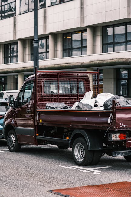 A dark red mini truck with a metal mesh safety barrier behind the cabin, parked on a city street in front of a modern office building with large windows and white facade panels. The truck's bed is loaded with various mixed waste items, including white and black garbage bags, some appearing partially open or disturbed, and other unidentifiable debris. The vehicle is positioned next to a white van, with a black lamppost visible nearby. The scene occurs during daylight with natural light illuminating the environment. The surrounding environment suggests an urban setting where private rubbish collection or on-site clearance services, such as those offered by Rubbish Collection Mayfair, can provide alternative waste disposal solutions for bulky waste removal.