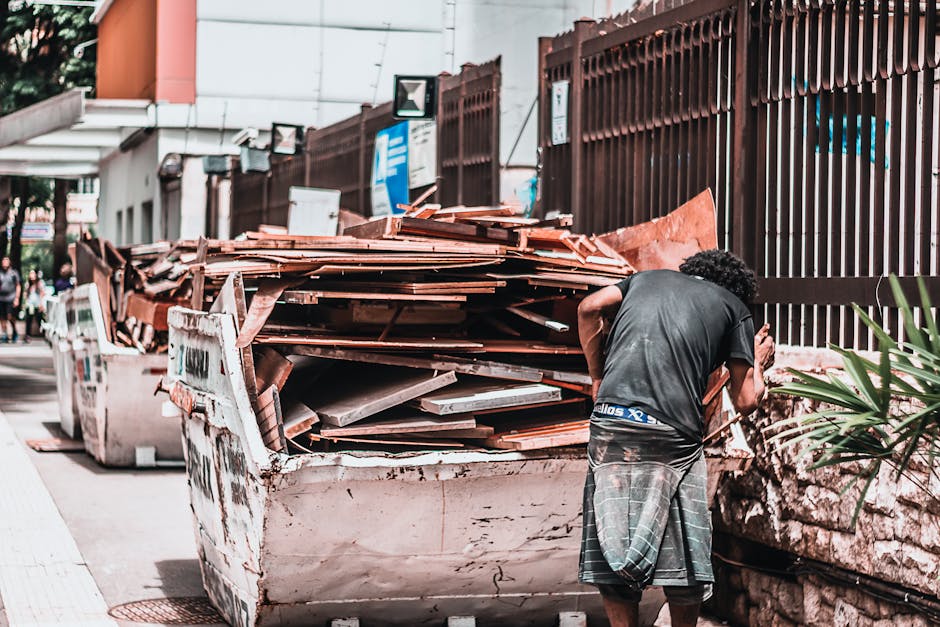 A man with curly black hair, wearing a dark T-shirt and striped shorts, is seen from behind while standing beside a large, white skip filled with a pile of burnt orange roof tiles, ceramic fragments, and assorted construction debris. The skip is positioned on a paved street, with additional skips and waste containers visible on the left side. In the background, a tall, dark metal fence runs along the edge of the property, and a modern building with a white facade and glass windows is partially visible above the fence. Small trees and plants are seen near the wall, adding some greenery to the scene. The environment appears to be an urban area, possibly during daylight hours, with natural light illuminating the scene evenly. This image offers a clear view of a private waste collection scenario, emphasizing on-site clearance activities that may be managed independently by waste removal specialists like Rubbish Collection Mayfair, supporting alternative rubbish disposal options outside of local authority services.