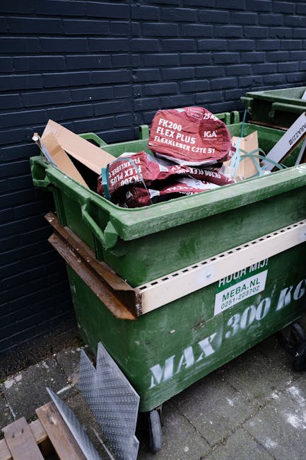 A large green wheelie bin filled with black plastic bags containing garden or landscaping waste, with red and white packaging labels visible on some bags. The bin is positioned on a paved surface next to a dark brick wall. The lid of the bin is open, revealing the contents, and a white plastic or metal support frame is attached to the side. The scene suggests an outdoor rubbish collection or waste disposal area, with a focus on domestic or commercial waste management. The environment appears tidy, with no visible litter outside the bin, and the setting is typical of a rear or side access point for waste removal services, possibly associated with private waste handling options like those offered by Rubbish Collection Mayfair.