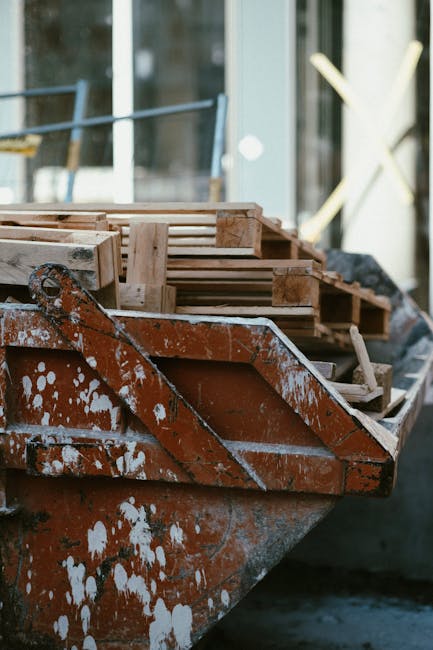 A man with curly black hair, wearing a dark T-shirt and striped shorts, is seen from behind while standing beside a large, white skip filled with a pile of burnt orange roof tiles, ceramic fragments, and assorted construction debris. The skip is positioned on a paved street, with additional skips and waste containers visible on the left side. In the background, a tall, dark metal fence runs along the edge of the property, and a modern building with a white facade and glass windows is partially visible above the fence. Small trees and plants are seen near the wall, adding some greenery to the scene. The environment appears to be an urban area, possibly during daylight hours, with natural light illuminating the scene evenly. This image offers a clear view of a private waste collection scenario, emphasizing on-site clearance activities that may be managed independently by waste removal specialists like Rubbish Collection Mayfair, supporting alternative rubbish disposal options outside of local authority services.