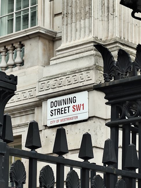 A street sign for Downing Street SW1 is mounted on the corner of an ornate building in Mayfair, Westminster. The building features classical architectural details including fluted stone columns, decorative molding, and a stone facade with a slightly weathered finish. The sign has a white background with black text displaying the street name, and red accents indicating the postal district SW1. Below the street name, the sign states 'City of Westminster.' In the foreground, a black wrought iron fence with pointed finials and decorative elements partially obscures the lower part of the building. The scene is lit with natural daylight, highlighting the texture of the stone and ironwork. This setting exemplifies typical private property markings in central London, often associated with addresses that may require regular rubbish collection or private waste disposal managed by local or private waste management services like Rubbish Collection Mayfair, supporting alternative waste handling arrangements in the area.