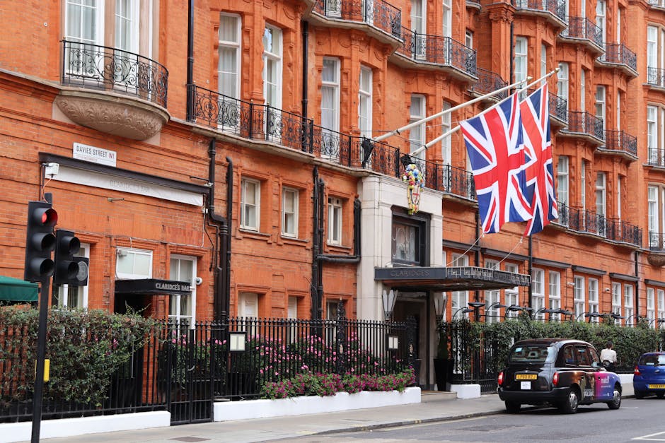 The image depicts a large, multi-storey red-brick building with classic Victorian architecture, featuring ornate stone balcony railings and neighboring windows with white frames. Two Union Jack flags hang prominently from poles extending from the building's facade, slightly fluttering in the breeze. In the foreground, a black iron fence encircles a small garden area with pink flowering plants and greenery, located along a sidewalk. A green awning marked 'Claridge's' covers the entrance to a nearby establishment, which appears to be part of the building. To the left, a traffic light displaying a red signal is visible, along with a white street sign reading 'Davies Street W1' attached to the building. A black taxi and a blue vehicle are parked along the street in front, with a pedestrian partially visible near the sidewalk. The overall scene offers an urban setting consistent with Mayfair's characteristic architecture and is relevant to private waste management and rubbish collection services operating in well-maintained city environments.