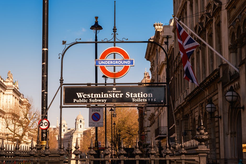 A street sign for Downing Street SW1 is mounted on the corner of an ornate building in Mayfair, Westminster. The building features classical architectural details including fluted stone columns, decorative molding, and a stone facade with a slightly weathered finish. The sign has a white background with black text displaying the street name, and red accents indicating the postal district SW1. Below the street name, the sign states 'City of Westminster.' In the foreground, a black wrought iron fence with pointed finials and decorative elements partially obscures the lower part of the building. The scene is lit with natural daylight, highlighting the texture of the stone and ironwork. This setting exemplifies typical private property markings in central London, often associated with addresses that may require regular rubbish collection or private waste disposal managed by local or private waste management services like Rubbish Collection Mayfair, supporting alternative waste handling arrangements in the area.